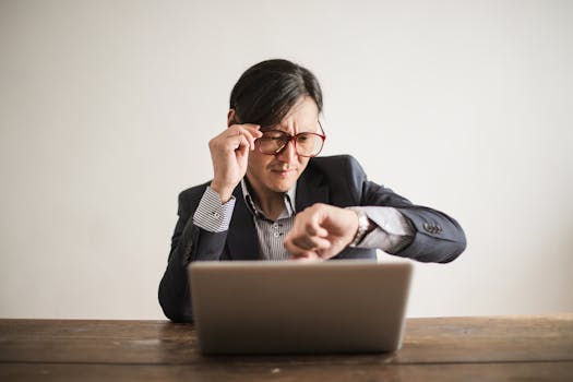 pexels-photo-3760810-3760810 Young frowning man in suit and glasses looking at wristwatch while waiting for appointment sitting at desk with laptop